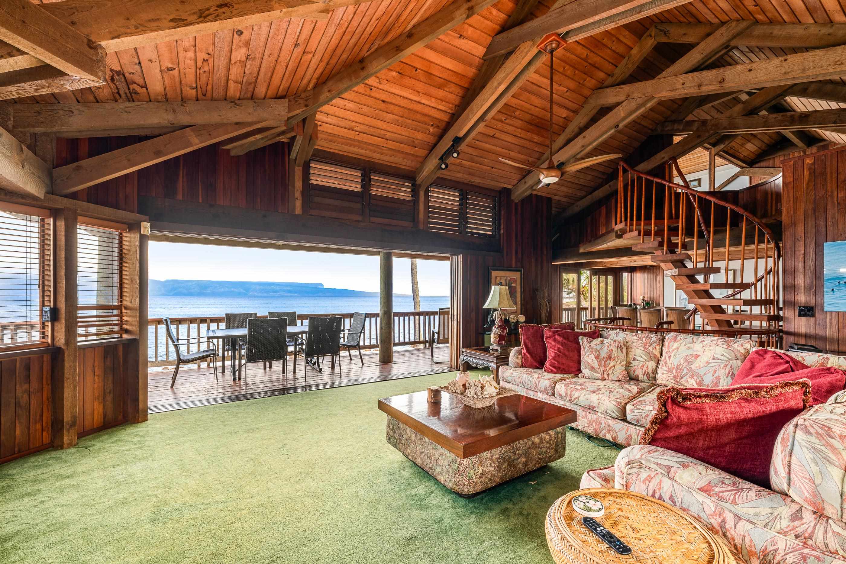 Living room under the vaulted timber ceiling: floral sofas on green carpet, the curved staircase rising to the loft, ocean through the windows