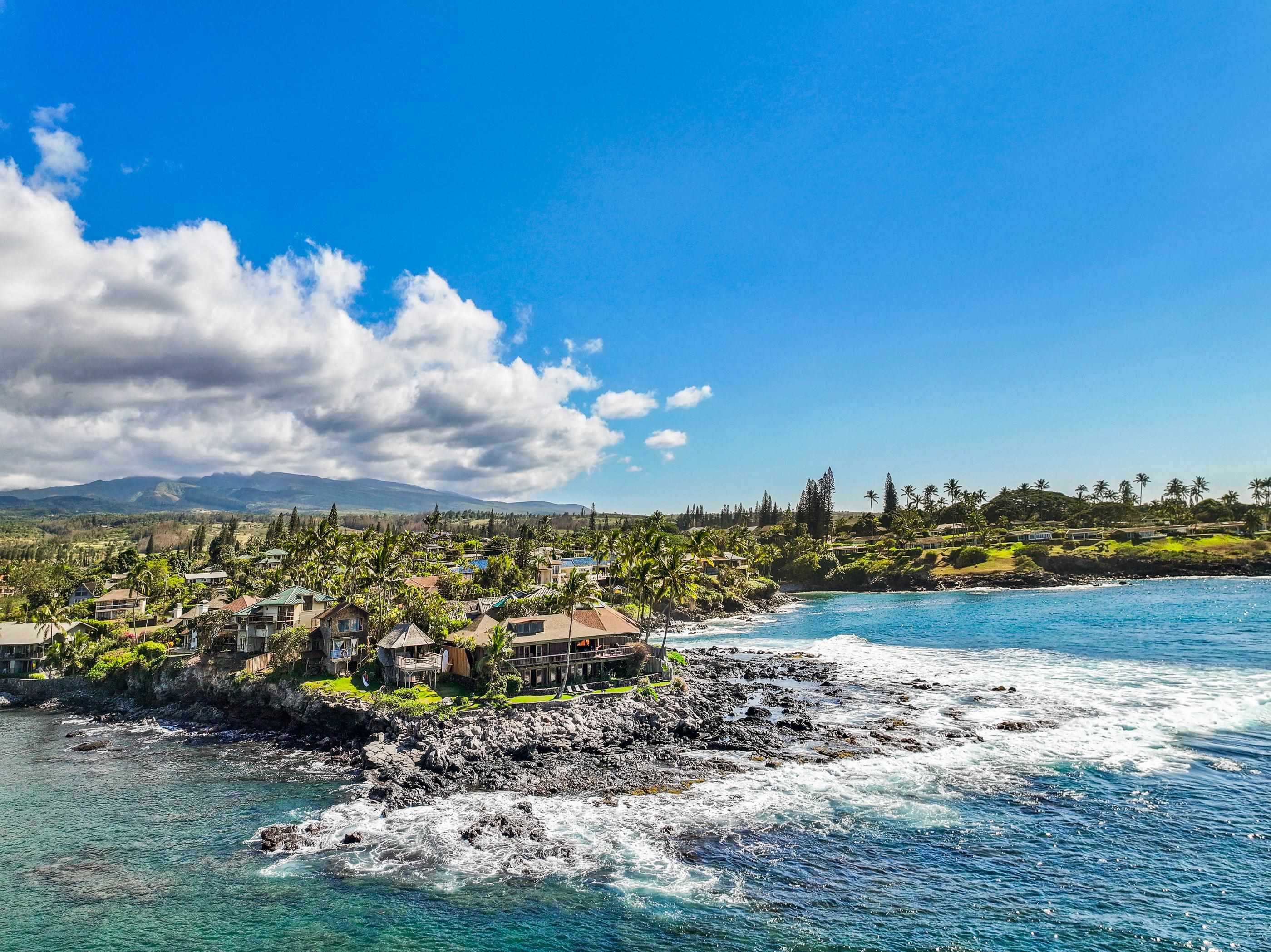 Wide aerial down the coast toward the West Maui mountains, surf breaking along the lava shore
